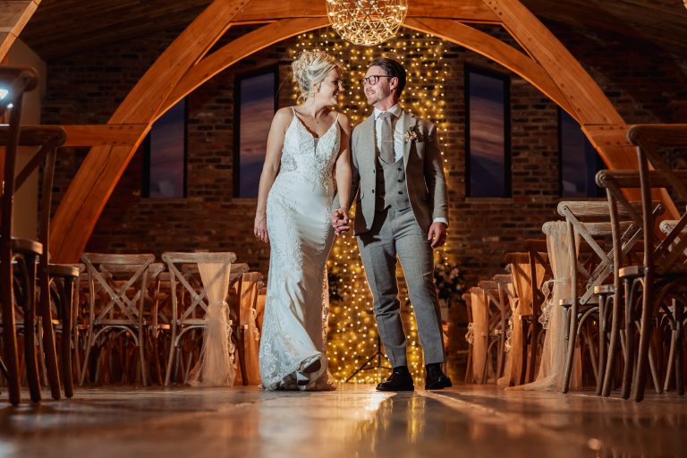 A bride and groom holding hands and smiling at each other as they walk down the aisle in Oakwood at Ryther, a beautifully decorated Yorkshire wedding venue with warm lighting and string lights glowing behind them.