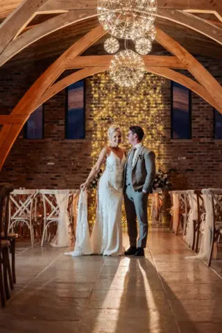 A bride and groom stand together in a rustic indoor venue at Oakwood at Ryther, with wooden arches, soft lighting, and string lights, smiling at each other on their Yorkshire wedding day.