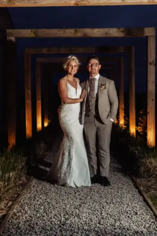 A bride in a white gown and a groom in a light grey suit stand arm in arm on a gravel path under wooden arches at Oakwood at Ryther, smiling at the camera, with warm lights illuminating this magical Yorkshire wedding night.