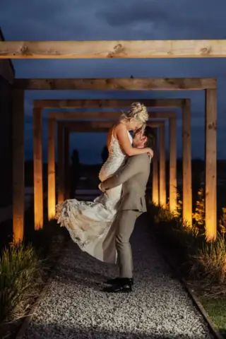 A groom in a gray suit lifts his bride in a white dress under wooden arches at night, with warm lights illuminating the pathway and romantic atmosphere—capturing the magic of an Oakwood at Ryther Yorkshire wedding.