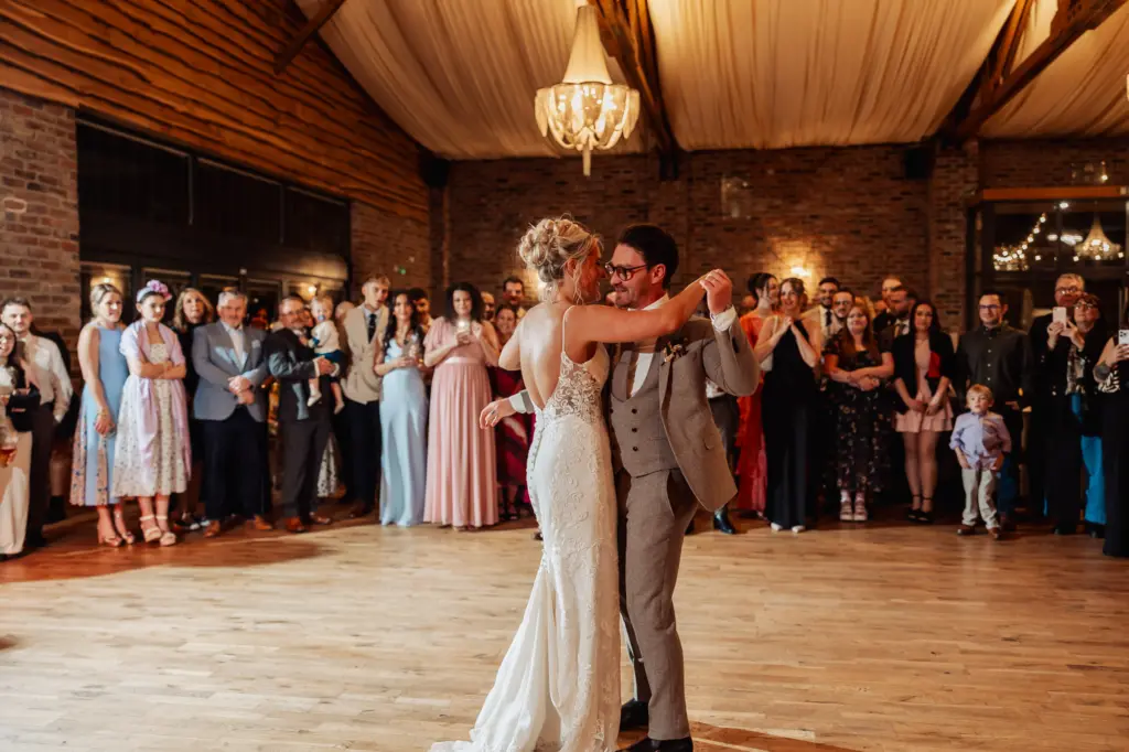 A bride and groom share their first dance in a rustic, warmly lit venue at Oakwood at Ryther, surrounded by smiling guests who watch and celebrate this unforgettable Yorkshire wedding moment.
