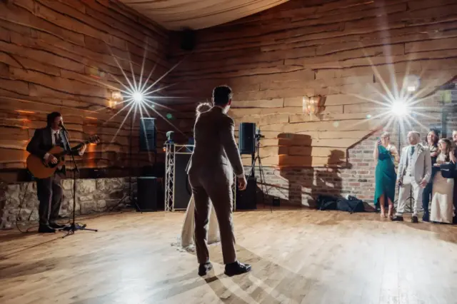 A couple dances together on a wooden floor in a warmly lit room at Oakwood at Ryther during a Yorkshire wedding, while a guitarist performs to the side. Several guests, dressed formally, watch and smile near the wall illuminated by bright spotlights.
