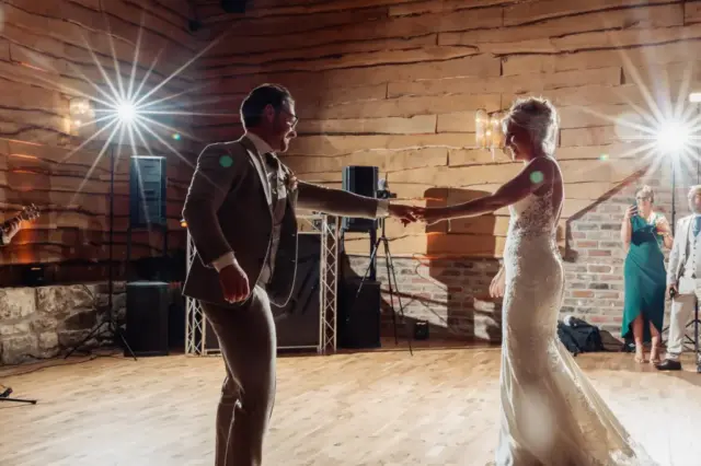 A bride and groom share a joyful first dance in the warmly lit Oakwood at Ryther, their smiles radiant. Wedding guests watch from the background as bright lights shine behind the happy couple at this stunning Yorkshire wedding venue.