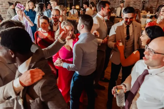A group of well-dressed people dance and socialize closely at a Yorkshire wedding, smiling and enjoying themselves in a warmly lit room with brick walls, capturing the joyful spirit of a celebration at Oakwood at Ryther.