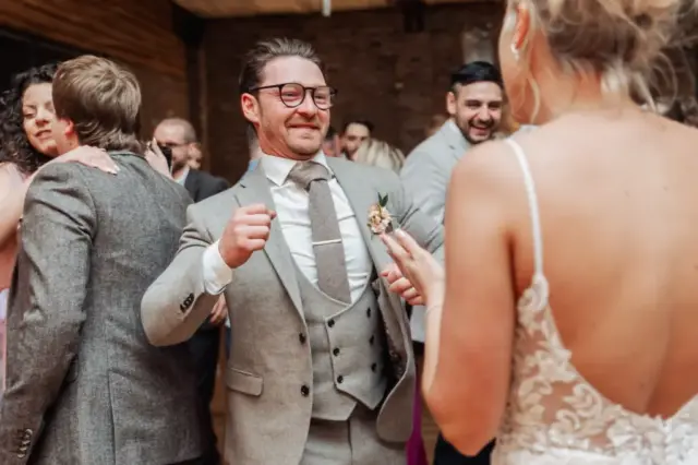 A man in a grey suit smiles and dances with a woman in a white lace dress at a lively Oakwood at Ryther Yorkshire wedding celebration, surrounded by other guests enjoying themselves.