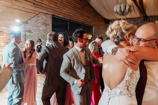 A group of people in formal attire dance and embrace at a Yorkshire wedding reception in a rustic venue with brick walls and wooden beams. A bride in a white dress hugs a man while others laugh and enjoy the celebration at Oakwood at Ryther.
