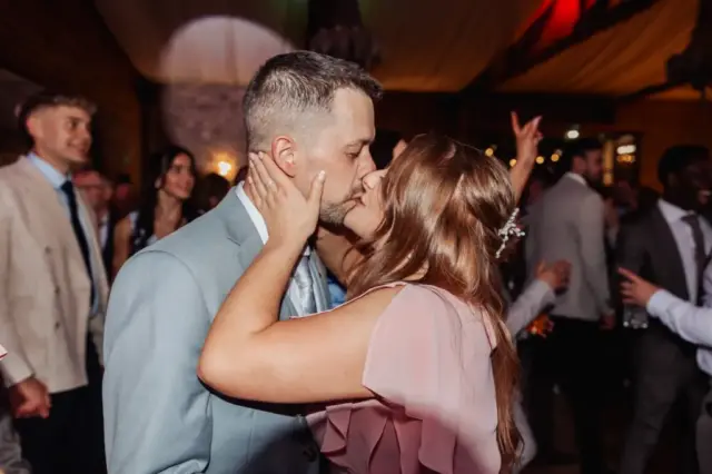 A couple shares a kiss on a crowded dance floor at an elegant Yorkshire wedding. The man wears a light blue suit and the woman wears a light pink dress, while other guests at Oakwood at Ryther celebrate in the background.