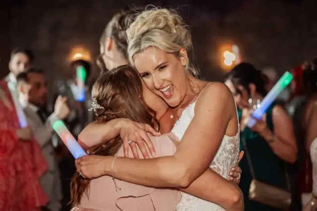 A smiling bride in a white dress warmly hugs a guest on a crowded Oakwood at Ryther dance floor, while people in the background hold colorful, glowing light sticks.