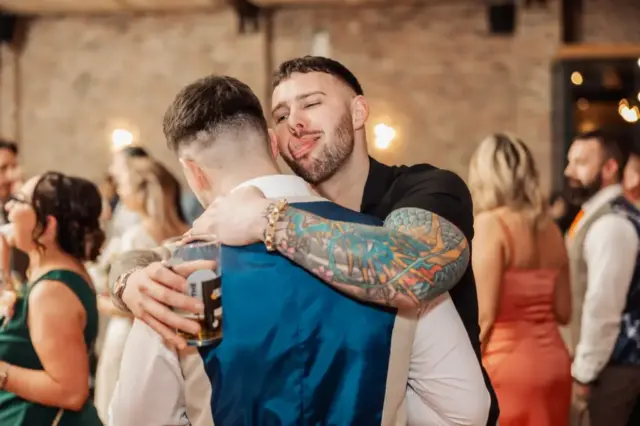 A tattooed man hugs another man in a blue vest while holding a beer, playfully sticking out his tongue. Several people mingle and laugh in the blurred background at this lively Yorkshire wedding indoor event.