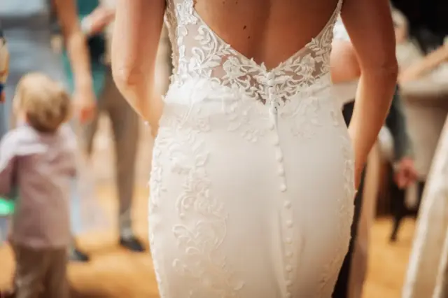 A close-up of a woman in a white lace wedding dress with buttons down the back, standing among blurred guests at an Oakwood at Ryther Yorkshire wedding reception.