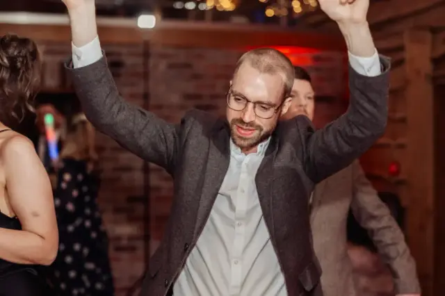 A man wearing glasses and a gray blazer smiles with his arms raised while dancing at a Yorkshire wedding at Oakwood at Ryther. Other guests are visible in the background, with exposed brick walls and warm lighting setting the scene.