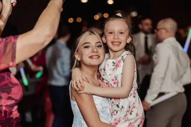 Two smiling girls, one older and one younger, pose together at a lively Yorkshire wedding. The older girl holds the younger in her arms as people in formal attire and glowing sticks fill the blurred background at Oakwood at Ryther.