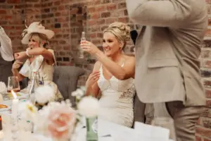 A smiling bride in a white dress raises a small glass for a toast at an Oakwood at Ryther Yorkshire wedding, seated beside another woman in a fancy hat. A man in a light suit stands nearby and the table is decorated with flowers and drinks.