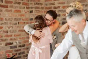 A man in glasses warmly hugs a woman in a pink dress with her back to the camera, while a smiling couple stands nearby against a brick wall—a joyful moment from an Oakwood at Ryther Yorkshire wedding.