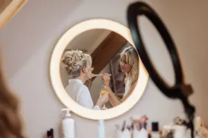 A woman with styled blonde hair is reflected in a round, illuminated mirror while a makeup artist applies her makeup before a Yorkshire wedding. She holds a glass of orange juice, with various beauty products on the table at Oakwood at Ryther.