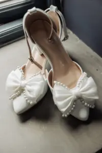 A pair of white, open-toe heels with ankle straps and large fabric bows decorated with pearls, perfect for a Yorkshire wedding, placed on a gray surface near a window.