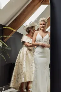 A bride in a white lace wedding dress stands in front of a mirror, smiling softly, as another woman in a cream dress and hat helps adjust her outfit at a modern Yorkshire wedding venue, Oakwood at Ryther, with sunlit wooden beams.