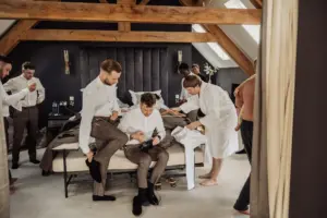 In a stylish Oakwood at Ryther bedroom, men prepare for a Yorkshire wedding—one is seated on the bed being assisted, others adjust shirts and ties, and one in a white robe; exposed beams and natural light fill the room.