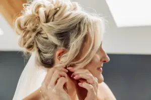 A bride with styled blonde hair and a veil smiles softly as she puts on an earring, preparing for her Yorkshire wedding at Oakwood at Ryther.