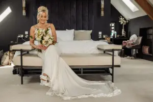 A smiling bride in a white wedding dress sits on a bench at the foot of a bed, holding her bouquet in an elegantly decorated bedroom with dark walls and modern lighting fixtures at Oakwood at Ryther, capturing the perfect Yorkshire wedding moment.