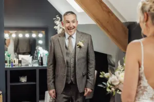 A man in a brown suit smiles widely at a woman in a bridal gown holding a bouquet. Indoors near a mirror with lights, the scene captures joyful Yorkshire wedding preparations, perhaps moments before their Oakwood at Ryther celebration.