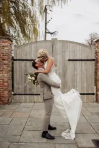 A groom in a light suit lifts and kisses his bride, who wears a white dress and holds a bouquet, in front of large wooden gates and brick pillars outdoors at Oakwood at Ryther during their Yorkshire wedding.