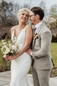 A bride in a white lace dress holding a bouquet smiles joyfully while her groom in a light gray suit and glasses stands beside her, holding her waist. They stand outdoors at Oakwood at Ryther, surrounded by lush greenery—a perfect Yorkshire wedding.