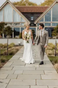 A bride in a white dress and groom in a light grey suit walk hand-in-hand outside on a stone pathway at Oakwood at Ryther, smiling at each other, with a modern building and greenery framing their Yorkshire wedding.