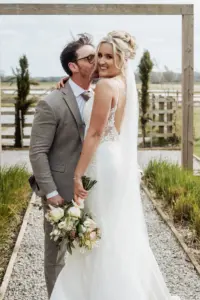 A groom in a grey suit kisses a smiling bride in a lace wedding dress holding a bouquet, standing together outdoors on a gravel path with greenery and wooden fencing at Oakwood at Ryther, the perfect Yorkshire wedding setting.