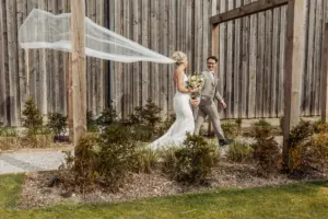 A bride in a white dress with a flowing veil and a groom in a tan suit hold hands and smile at each other while walking outdoors near a wooden fence and garden at their beautiful Oakwood at Ryther Yorkshire wedding.