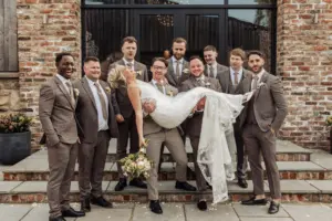 A joyful bride in a white gown is being lifted by several groomsmen in tan suits, smiling and celebrating outside Oakwood at Ryther—a stunning Yorkshire wedding venue with a rustic brick building and large windows.