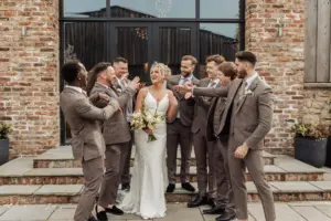 A bride in a white dress stands smiling with a bouquet as six groomsmen in brown suits surround her, laughing and pointing at her, in front of the rustic Oakwood at Ryther, capturing a joyful Yorkshire wedding moment.