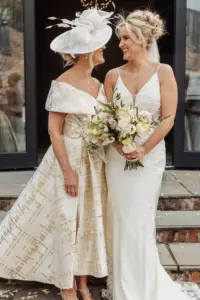 A bride in a white lace gown holding a bouquet stands smiling next to a woman in an off-shoulder dress with a large white hat. They share a joyful moment outside Oakwood at Ryther, capturing the spirit of a beautiful Yorkshire wedding.