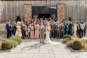 A large wedding group poses outdoors at Oakwood at Ryther in front of a rustic barn. The bride and groom stand at the front, surrounded by family and friends dressed in formal attire, smiling for this Yorkshire wedding photo.