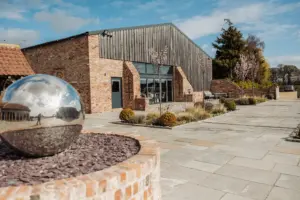A modern barn-style building with brick walls and large glass doors at Oakwood at Ryther, surrounded by landscaped gardens and a paved courtyard—perfect for a Yorkshire wedding. A reflective silver sphere sculpture sits in the foreground under a partly cloudy sky.