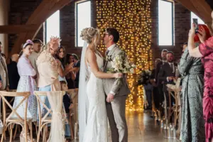A bride and groom share a kiss at their Oakwood at Ryther wedding, standing at the aisle’s end as guests clap. Warm fairy lights hang behind them, creating a festive, romantic atmosphere perfect for a Yorkshire wedding celebration.