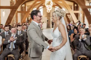 A bride and groom smile at each other and hold hands during their wedding ceremony at Oakwood at Ryther, a rustic Yorkshire wedding venue, as guests seated behind them clap and watch joyfully.