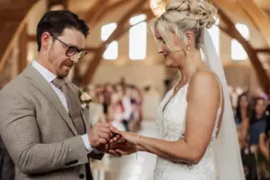 A groom gently places a ring on the bride’s finger during a Yorkshire wedding ceremony at Oakwood at Ryther, in a bright venue with guests seated in the background. The couple looks at each other lovingly.