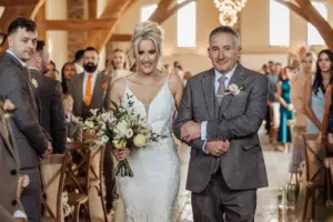 A smiling bride in a white dress holds a bouquet as she walks down the aisle arm-in-arm with an older man in a grey suit, surrounded by guests in the bright, rustic Oakwood at Ryther Yorkshire wedding venue.