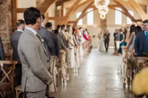 A man in a suit stands at the front of an aisle, looking toward guests who rise as someone walks down the aisle at Oakwood at Ryther, a rustic, sunlit Yorkshire wedding venue beautifully decorated for the occasion.
