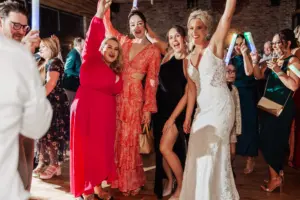 A group of people, including a woman in a wedding dress and others in colorful dresses, smile and pose with raised arms while dancing at a lively indoor Yorkshire wedding celebration at Oakwood at Ryther.