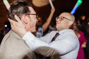 Two men in dress shirts and glasses smile and embrace at a lively Oakwood at Ryther wedding, with people and colorful lights blurred in the background.