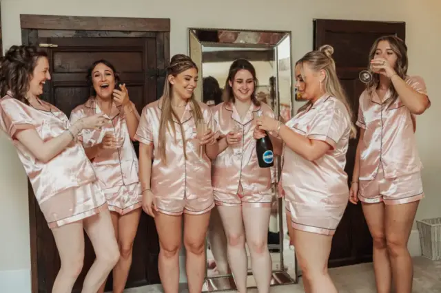 Six women in matching pink satin pajamas stand together indoors at Eyam Hall, smiling and laughing. One opens a bottle of sparkling wine while others hold glasses, celebrating weddings. A mirror and dark wooden doors are in the background.