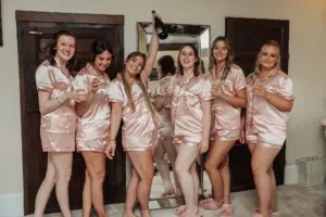 Six women in matching pink satin pajamas and slippers stand indoors at Eyam Hall, smiling and holding glasses of champagne. One woman raises a bottle in celebration—they appear joyful and ready for weddings or a special occasion.