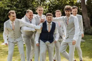 A group of seven men, dressed in light grey suits with pale pink ties, laugh and playfully gather around a man in a navy blue vest outdoors at Eyam Hall on a sunny day. They appear to be celebrating weddings at this charming venue.
