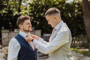 Two men in dress shirts and vests stand outdoors at Eyam Hall; one adjusts the others tie while both smile, capturing a joyful moment often seen at weddings. Trees and garden furniture fill the sunlit background.