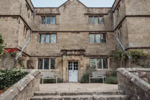 A historic stone building at Eyam Hall with symmetrical windows, a white door, and two wooden benches in front, surrounded by flowers and greenery—an ideal setting for romantic weddings.