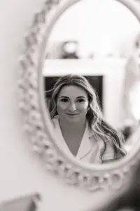 Black and white photo of a woman with long hair and a slight smile, reflected in an ornate oval mirror at Eyam Hall; the softly blurred background hints at the charm of weddings held in this elegant setting.