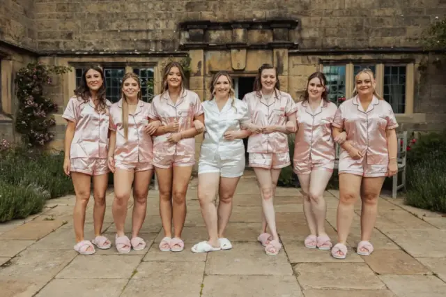 Seven women stand arm-in-arm outdoors in matching short-sleeved pajamas and slippers, smiling in front of Eyam Hall with greenery. Six wear pink, one wears white—capturing a joyful, pre-wedding moment at this historic weddings venue.