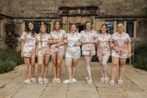Seven women stand arm-in-arm outdoors in matching short-sleeved pajamas and slippers, smiling in front of Eyam Hall with greenery. Six wear pink, one wears white—capturing a joyful, pre-wedding moment at this historic weddings venue.